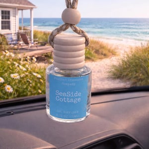 Può includere: Un diffusore di olio in vetro trasparente con un tappo in legno e un gancio in corda. L'etichetta recita "SeaSide Cottage" e "Oil Diffuser 0.33 oz / 10ml". Lo sfondo mostra una scena di spiaggia con un cottage e l'oceano.