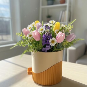 Puede incluir: Un arreglo floral en una maceta crema y naranja. El ramo incluye tulipanes rosas, amarillos y morados, margaritas blancas e jacintos morados, con follaje verde. La maceta tiene un diseño curvo y un asa pequeña.