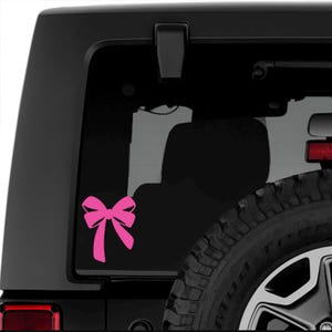 Peut inclure: Autocollant en forme de nœud rose sur la lunette arrière d'une Jeep noire. Le nœud est un design simple avec deux boucles et deux queues.