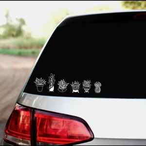 May include: A white car with a black rear window. A white vinyl decal of potted plants is on the rear window. The decal features seven potted plants with different types of leaves.
