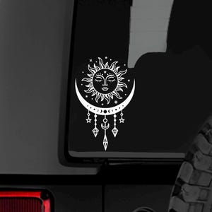 May include: White celestial-themed decal featuring a sun with a face, a crescent moon, stars, and dangling geometric shapes. The design is a detailed illustration, suitable for car windows or other smooth surfaces.