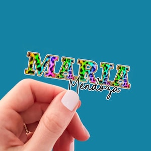May include: A colorful sticker with the name "MARIA Mendoza" in a vibrant leopard print design. The name "MARIA" is in bold, block letters, while "Mendoza" is in a cursive font. The sticker is held against a teal background.