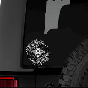 May include: White vinyl decal of a bee in a honeycomb with flowers surrounding it. The decal is on a black car.