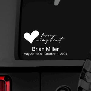 Puede incluir: Un adhesivo de vinilo blanco sobre una superficie oscura que presenta un icono de corazón y el texto en cursiva "forever in my heart". Debajo, el nombre "Brian Miller" en una fuente sans-serif, seguido de las fechas "May 20, 1990 - October 1, 2024".