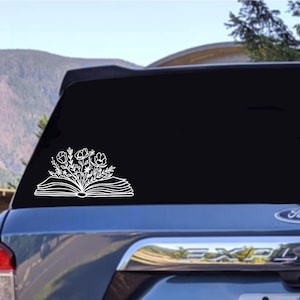 Puede incluir: Una calcomanía de vinilo blanco de un libro abierto con flores que salen de él. La calcomanía está en la ventana trasera de un coche azul oscuro.