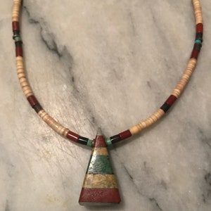 May include: A necklace made of beige, red, and black beads with a triangular pendant made of layered green, brown, and red stones.