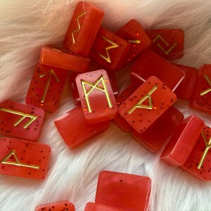 May include: A collection of red resin runes with gold-coloured symbols and glitter. The runes are rectangular and vary in shape. The background is a soft, white, fluffy surface.