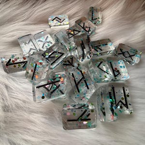 Puede incluir: Una colección de piedras rúnicas de resina transparente con símbolos negros y purpurina colorida. Las runas están dispersas sobre una superficie blanca y esponjosa. Cada piedra tiene una forma y tamaño diferentes.