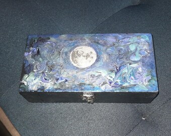 Puede incluir: Una caja de madera con acabado negro y un diseño de luna plateada en la tapa. La caja tiene un patrón de mármol azul, verde y plateado en la tapa.