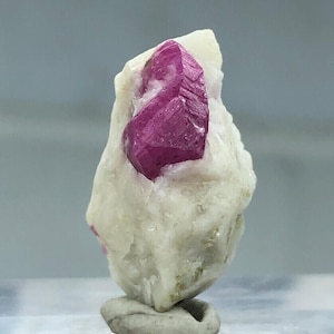 May include: A pink ruby crystal embedded in a white matrix. The crystal is faceted and has a sharp point. The matrix is rough and textured.