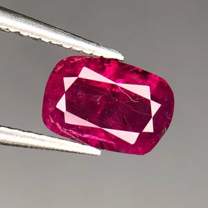 May include: A deep red gemstone with a faceted cut. The gemstone is held in tweezers against a gray background.