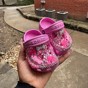 Pode incluir: Um par de sapatos Crocs rosa, ricamente adornados com strass, laços e detalhes em forma de coração. A palavra "Barbie" é visível em um sapato. Os sapatos são segurados na mão de uma pessoa.