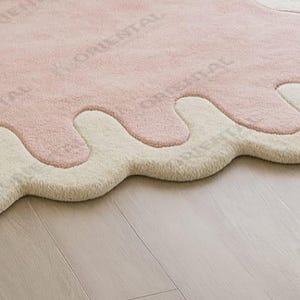 Modern abstract scalloped edge pink rug in size 8x10 ft, styled with white bedding, natural wood flooring and indoor greenery Luxurious hand tufted blush pink rug with cream wave border, 6x9 ft, styled in a cozy minimalist bedroom with natural light and soft tones.