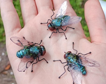 May include: Three small insect figurines resembling houseflies, each featuring a metallic iridescent blue and green body, dark red eyes, and translucent, veined wings. The legs are made of thin, dark, rigid wire extending from the thorax.