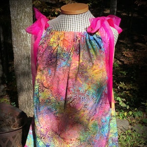 May include: A colorful, tie-dye patterned dress with pink ribbon straps and a white crochet trim. The dress is on a mannequin and has a brown and white checkered background.