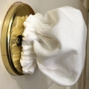 Könnte beinhalten: Eine weiße Stoff-Türklinkenabdeckung ist um einen Messing-Türknauf gewickelt. Die Abdeckung besteht aus einem weichen, strukturierten Material und ist am Boden gerafft. Der Türknauf ist an einer weißen Tür befestigt.