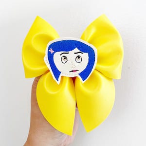 May include: A large, bright yellow hair bow with a cartoon character face in the center. The character has blue hair, white skin, and large eyes. The bow is held in a person's hand against a white background.