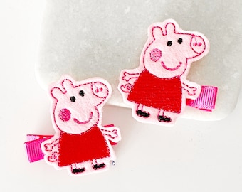 May include: Two pink pig hair clips with red dresses and black shoes. The clips are attached to pink ribbon.