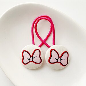 May include: Two white fabric hair ties featuring a pink bow design with a red outline. The bows are attached to a bright pink elastic band. The hair accessories are displayed on a white, oval-shaped dish.