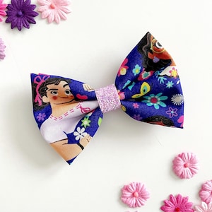 May include: A fabric hair bow featuring a character print on a blue background with floral and butterfly accents. The bow's center is a glittery light purple. Decorative paper flowers in shades of pink and purple surround the bow.