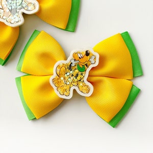 May include: Yellow and green hair bows with a cartoon Pluto character embellishment. The bows are made of a bright yellow ribbon with green edges. The Pluto character is in a clear, glitter-filled charm.
