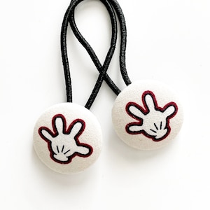 May include: Two white fabric-covered hair ties with black elastic bands. Each tie features a cartoon hand design with a red outline and black details. The hair accessories are set against a white background.