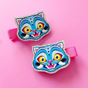 May include: Two blue cat-faced hair clips with glitter trim and pink ribbon. The cat faces have yellow eyes, red mouths, and white teeth. The background is a solid pink.
