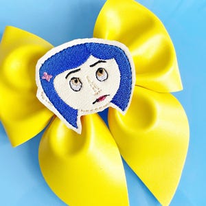 May include: A bright yellow hair bow with a cartoon character face in the center. The character has blue hair, a pink bow, and a worried expression. The bow is made of a shiny material and is set against a light blue background.