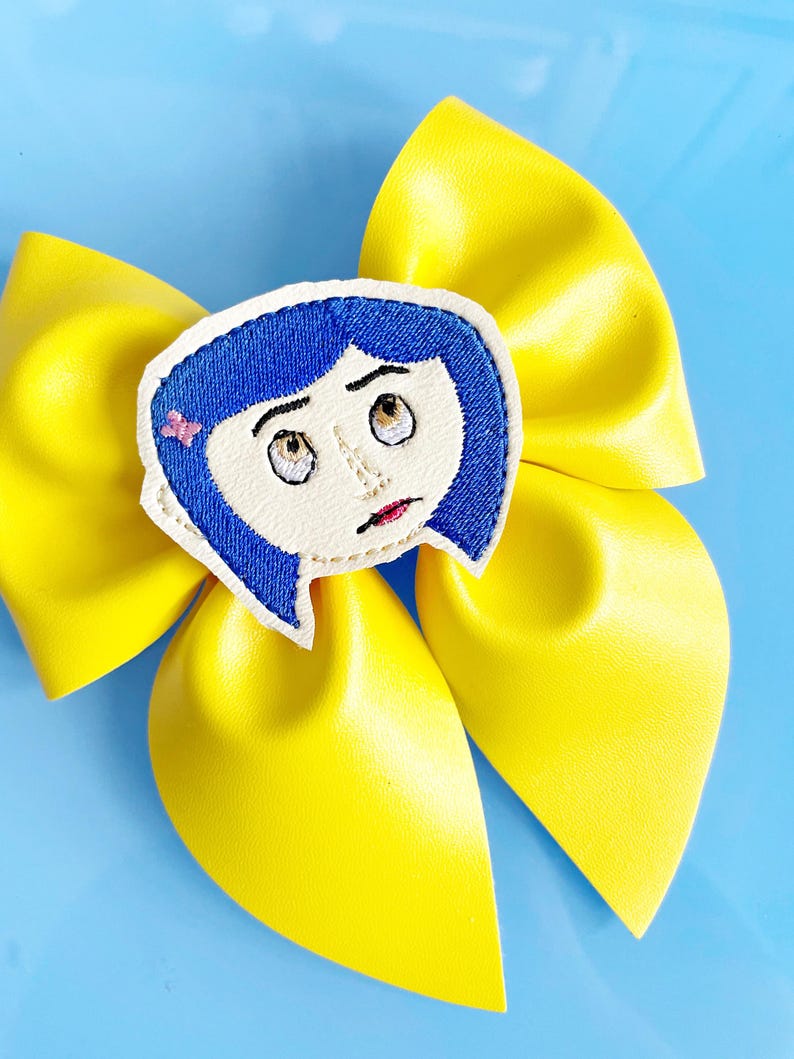 May include: A bright yellow hair bow with a cartoon character face in the center. The character has blue hair, a pink bow, and a worried expression. The bow is made of a shiny material and is set against a light blue background.
