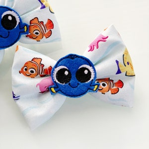 Può includere: Due fiocchi per capelli in tessuto bianco con un'applicazione di personaggio di pesce blu. Il tessuto ha uno sfondo bianco con un motivo ripetitivo di pesci colorati.