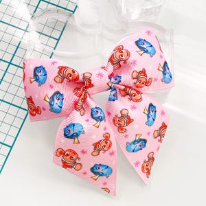 Peut inclure: Un nœud pour cheveux rose avec un motif répété de poissons de dessin animé, dont Dory et Nemo. Le nœud est fait d'un tissu doux et texturé.