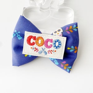 May include: A blue fabric bow tie with a colourful floral pattern and the word "COCO" embroidered on a white patch. The bow tie is attached to a clear plastic form with a white ribbon.