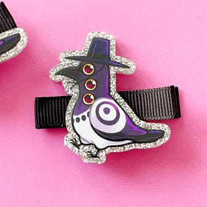 May include: A decorative hair clip featuring a purple pigeon wearing a black hat with three red eyes. The bird has a white chest and a swirling design on its wing. The clip is outlined with silver glitter and attached to a black ribbon.