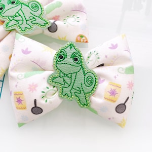 May include: Two white fabric hair bows with a green embroidered chameleon design. The fabric has a light pink, yellow, and green pattern with stars and other small designs.