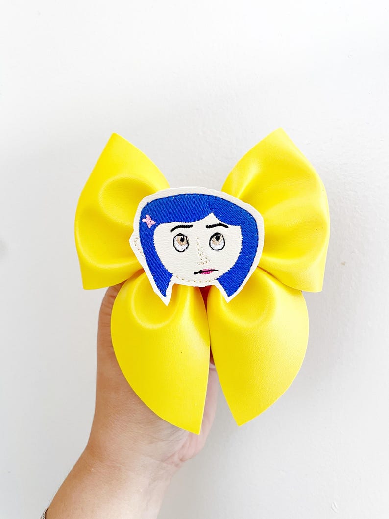 May include: A large, bright yellow hair bow with a cartoon character face in the center. The character has blue hair, white skin, and large eyes. The bow is held in a person's hand against a white background.