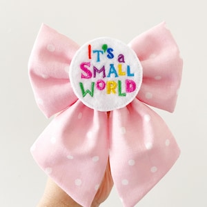 May include: A pink polka dot bow with a white circular center. The center has the embroidered text "It's a SMALL WORLD" in various colors. The bow is made of fabric and is likely for hair accessories.