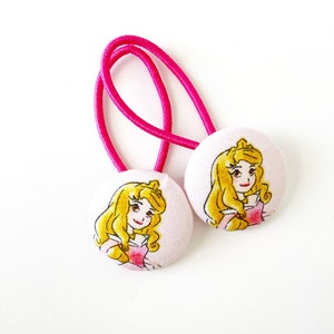 May include: Two pink hair ties with white fabric buttons featuring a cartoon image of Princess Aurora from Sleeping Beauty.