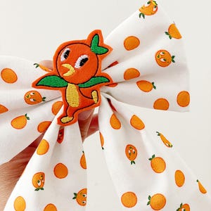 May include: A white fabric bow with a repeating pattern of orange cartoon oranges and orange polka dots. An orange bird applique with green wings and a yellow belly is centered on the bow. The bow is held by a hand.