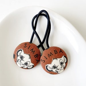 Puede incluir: Dos coleteros marrones de Simba con bandas elásticas negras. Cada uno presenta un diseño circular de tela con la palabra "SIMBA" y una ilustración de cachorro de león de dibujos animados. Los coleteros se muestran sobre una superficie blanca curvada.