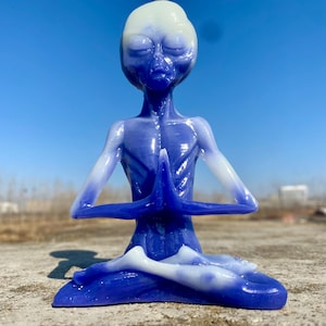 May include: A blue and white alien figurine in a seated yoga pose with hands together in prayer. The figurine is a gradient of blue and white, with a smooth, glossy finish. The background is a clear blue sky.