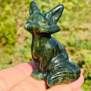 May include: A dark green fox figurine carved from stone. The fox is in a seated position with its ears perked up and tail curled. The figurine is held in a hand against a blurred green background.