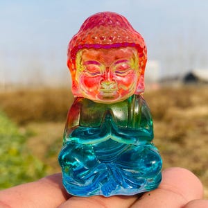 Puede incluir: Una pequeña figurita de vidrio de colores de un Buda meditando. La figurita está hecha de vidrio transparente con capas de rojo, naranja, amarillo, verde y azul.