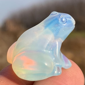 Puede incluir: Una pequeña figura de rana de ópalo blanco translúcido con matices iridiscentes azules y rosados. La rana está sentada con las patas metidas debajo del cuerpo.