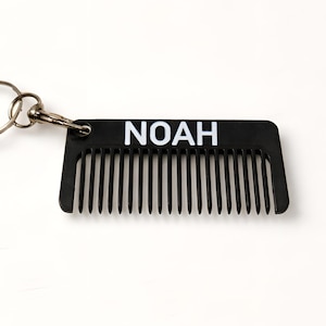 May include: A black comb-shaped keyring with the name "NOAH" in white block letters. The comb has fine teeth and is attached to a silver key ring. The keyring is on a white background.