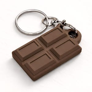 May include: A brown chocolate bar keyring with a silver key ring and clasp. The keyring is a rectangular shape with four embossed squares, resembling a chocolate bar. The background is white.