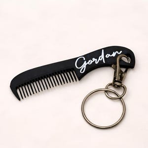 May include: A black comb keychain with the name "Gordan" in white script. The comb has fine teeth and a curved handle. It is attached to a metal keyring and clasp.