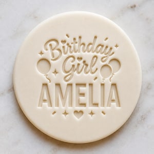 May include: A round, off-white cookie stamp featuring the embossed text "Birthday Girl" in a cursive script, the name "AMELIA" in large block capital letters, and decorative icons including two balloons, several small stars, and a heart.