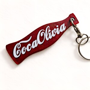 May include: A red, bottle-shaped keyring with the text "Coca Olivia" in white script. The keyring has a silver-coloured clasp and ring. The background is white.