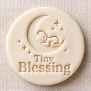 May include: A circular off-white stamp featuring an embossed design of a baby sleeping on a crescent moon surrounded by small stars. The text "Tiny Blessing" is debossed in a clear serif font below the moon graphic on the flat surface.