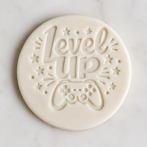May include: A round, off-white cookie with the words "Level Up" and a game controller design etched into the surface. Stars and radiating lines surround the text and controller, creating a celebratory feel. A fun treat for gamers.